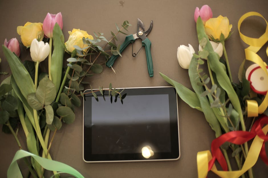 A flat-lay arrangement of fresh tulips and yellow roses with green stems and leaves, partially wrapped with colorful ribbons, positioned around a tablet device on a neutral surface. A pair of pruning 