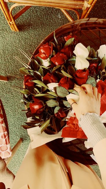 A floral arrangement featuring a bouquet of fresh red and white roses with green foliage, wrapped in light beige paper, and arranged on a dark wooden table. The bouquet showcases vibrant, healthy peta