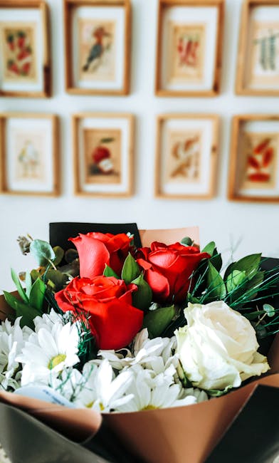 A close-up of a floral bouquet featuring vibrant red roses with velvety petals and fresh white blooms, possibly chrysanthemums or daisies, arranged with lush green foliage. The bouquet is wrapped in p