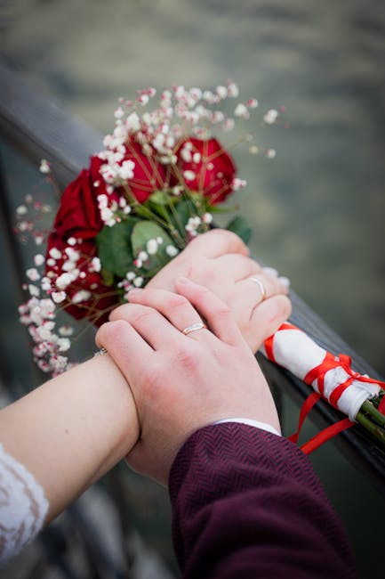 A close-up of a bride and groom's hands resting on a railing, with the bride holding a bouquet of deep red roses accented by small white baby's breath flowers. The bouquet is wrapped in white paper wi