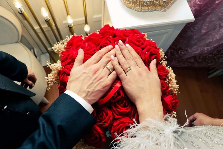 A close-up view of a newlywed couple's hands, both wearing wedding rings, resting atop a large, round bouquet of red roses interspersed with white baby's breath. The bouquet is lush and vibrant, with