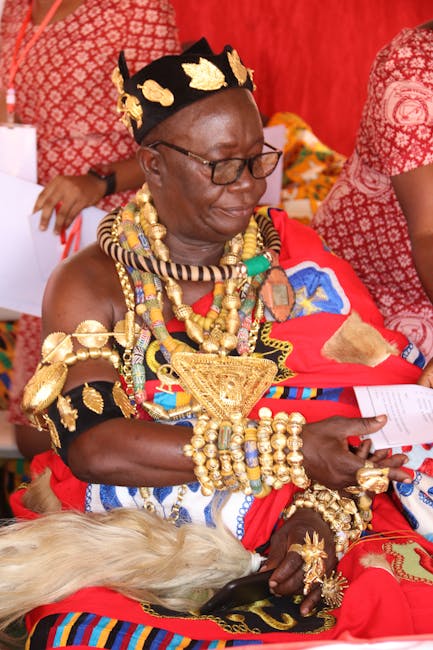 The image features an elderly woman dressed in vibrant traditional African attire, adorned with an elaborate array of gold jewelry, including necklaces, bracelets, earrings, and a crown-like headpiece