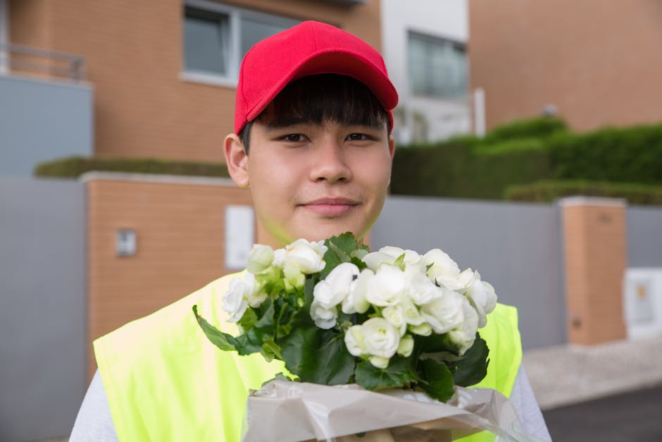 A young male flower delivery courier wearing a red cap and a neon yellow safety vest is standing outdoors in front of modern houses with brick and concrete walls. He is smiling gently while holding a 