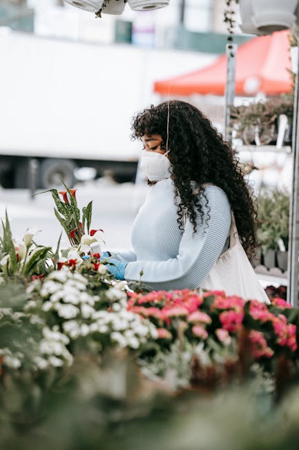 A woman with curly dark hair, wearing a white face mask and a light grey sweater, stands at an outdoor flower market stall. She is carefully arranging a bouquet of fresh red and white flowers, includi
