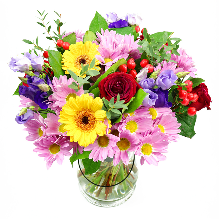 This abundant bouquet, displayed in a simple clear glass vase against a bright white background, feels both polished and inviting, like a thoughtful gift arriving at a doorstep in Gants Hill. At its core, velvety red roses create a rich, romantic anchor, their layered petals forming intricate spirals that draw the eye inward. Nearby, bold yellow gerbera daisies with dark, warm centres offer a burst of sunshine colour, their flat, daisy-like faces bringing a touch of modern flair. Surrounding these are plush clusters of pink and soft lilac daisies or chrysanthemums, whose countless slender petals and neat yellow centres add both volume and a gentle, diffused glow, softening the contrast of the brighter blooms. Threaded among them, lisianthus flowers in shades of purple and lavender stretch gracefully upward, their bell-shaped blossoms and lightly ruffled edges introducing a cool, elegant note that balances the warm reds and yellows. Between the petals, bright red hypericum berries appear in rounded clusters, smooth to the eye and adding playful texture. The whole arrangement is framed by varied greenery: sturdy, dark leaves mingle with finer, lighter sprigs to suggest fresh growth and movement, reminiscent of the greenery found on walks between Gants Hill and nearby Cranbrook Road. Through the clear sides of the vase, you can see the carefully cut stems, neatly fanned in clear water, a sign of the florist's practiced hand and attention to freshness. The overall effect is one of cheerful generosity and subtle sophistication, ideal for marking a special occasion, welcoming someone to a new home in Ilford, or simply bringing a lively splash of garden colour indoors.